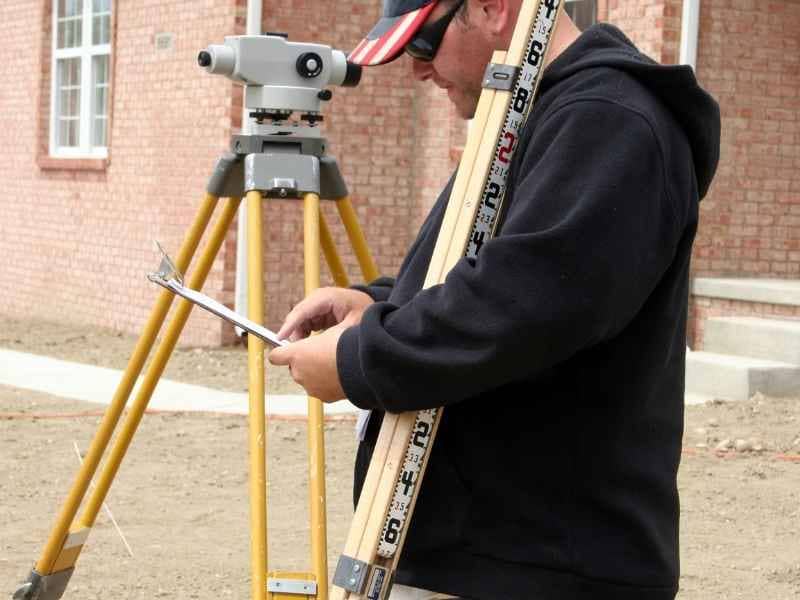Boundary Line Surveys St Louis Missouri | St. Louis Land Surveying Surveyor in a black hoodie holds a clipboard beside a wooden measuring rod and a tripod-mounted surveying instrument at a construction site with a brick building in the background.